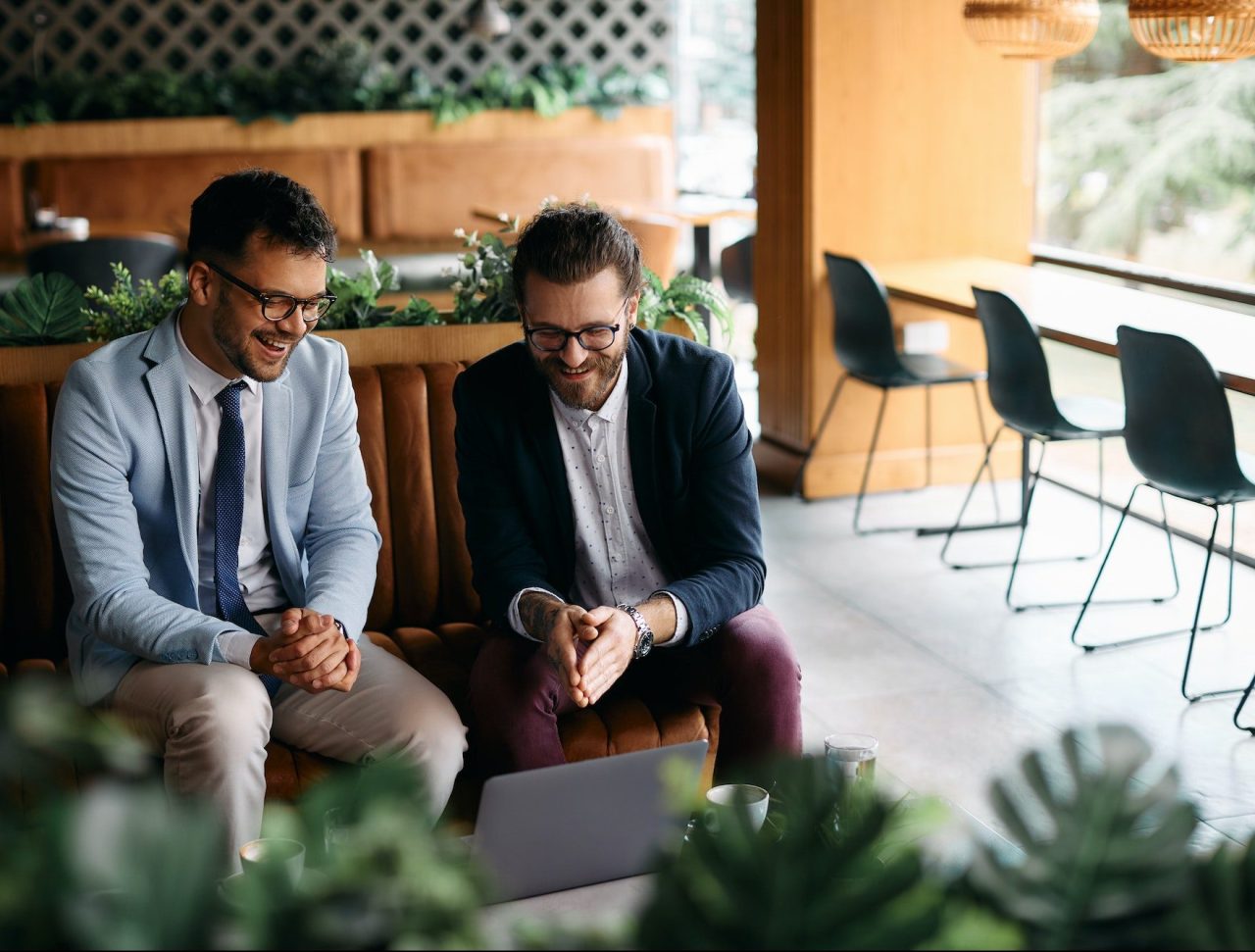 cheerful businessmen having fun while using laptop in a cafe e1689501076718